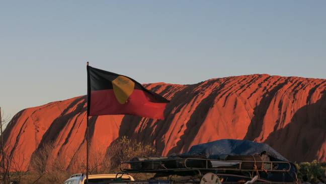 原住民的旗帜在澳大利亚中部的乌鲁鲁鲁（Uluruu）或艾尔斯（Ayers）岩石前飞。砂岩巨石对澳大利亚土著人是神圣的。学分：Leigh Henningham / Alamy。