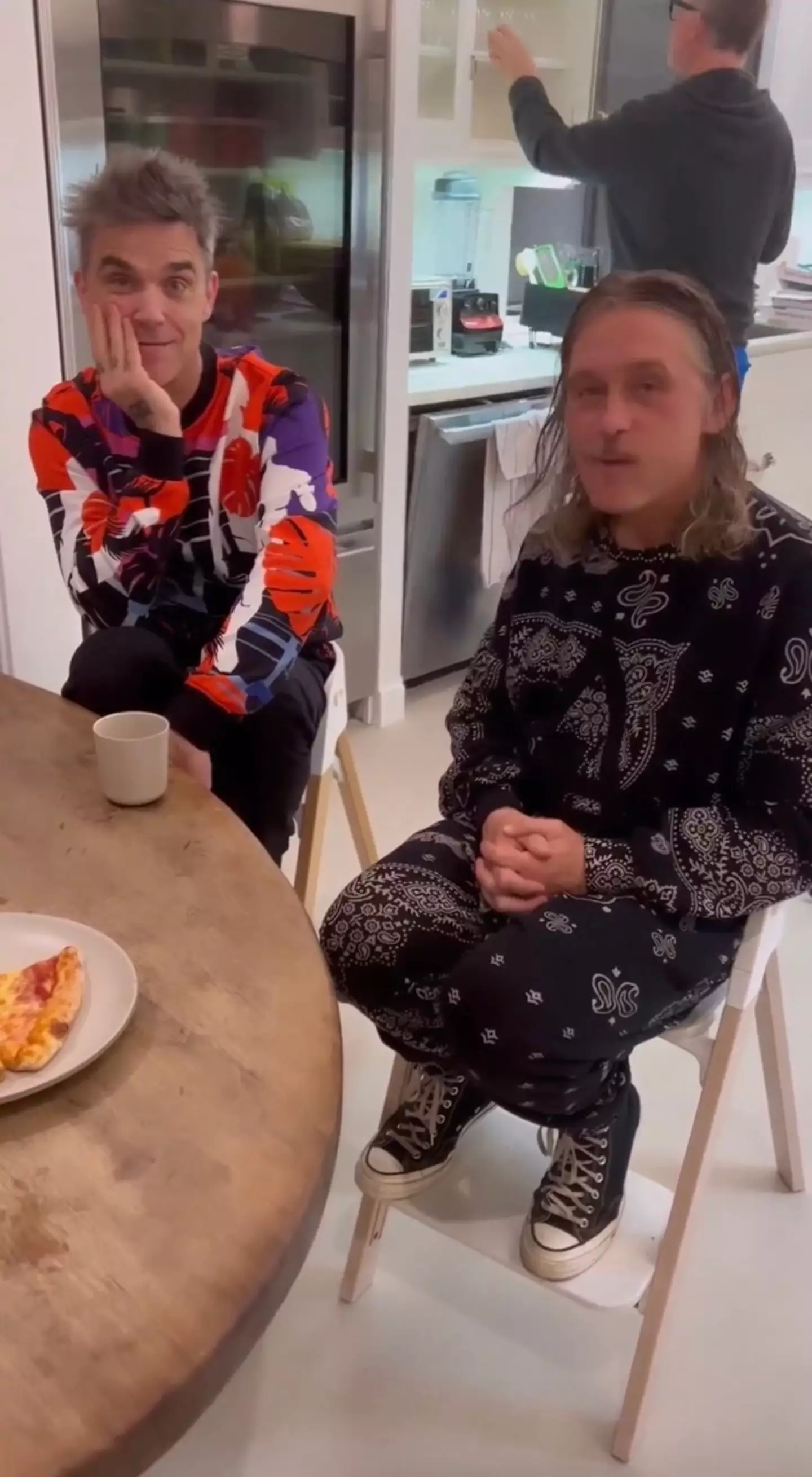 Robbie, 48, and Mark, 50, are both randomly tucking into a slice of pizza while sat in 'baby' high chairs.