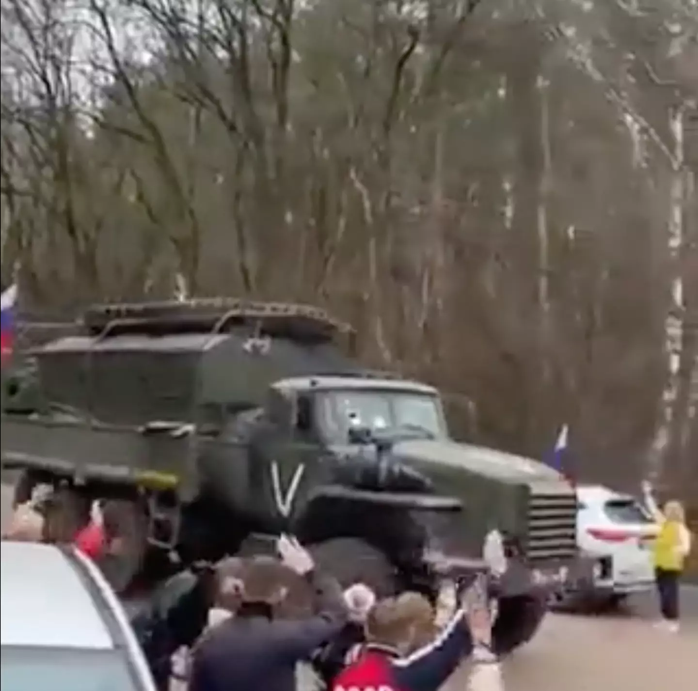 A Russian military convoy with a white painted symbol on its side, being waved into battle by passersby.