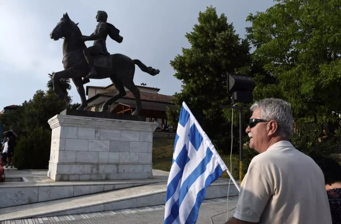 A man holds a Greek flag next to a statue of Alexander the Great.